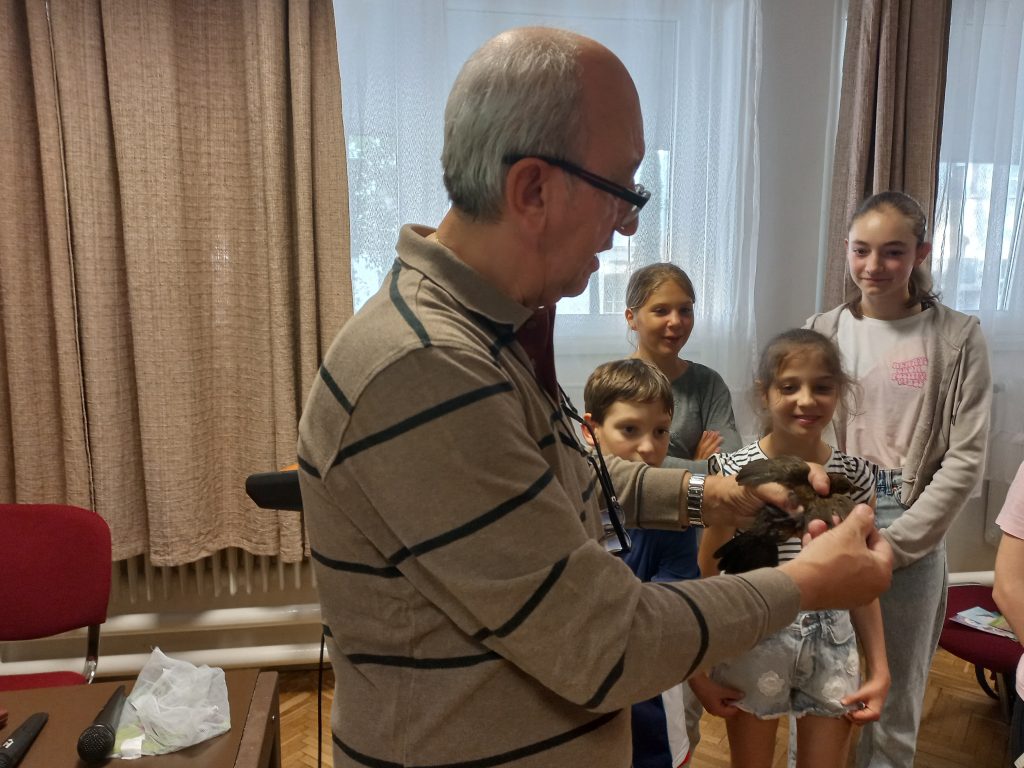 Dr. Kis Ernő kezében tartva mutatja a közönségnek a meggyűrűzött fekete rigót. A háttérben gyerekek csoportja mosolyogva figyeli a madarat.