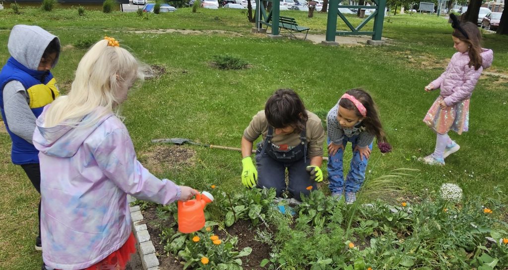 A gyerekek kis, színes locsolókannákból öntözik a virágokat, a főkertész az ágyás szélénél térdelve segít nekik